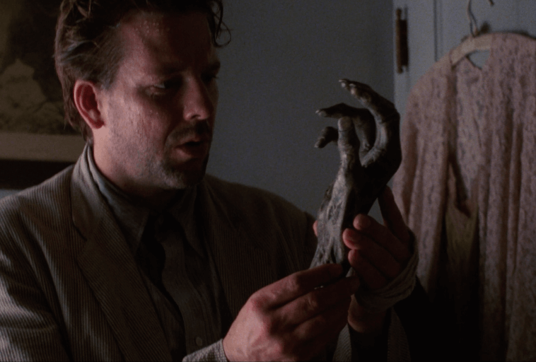 Mężczyzna w pasiastej kurtce przygląda się z niepokojem ciemnej, szponiastej, zmumifikowanej dłoni, stojąc w pomieszczeniu obok płaszcza wiszącego na haczyku.