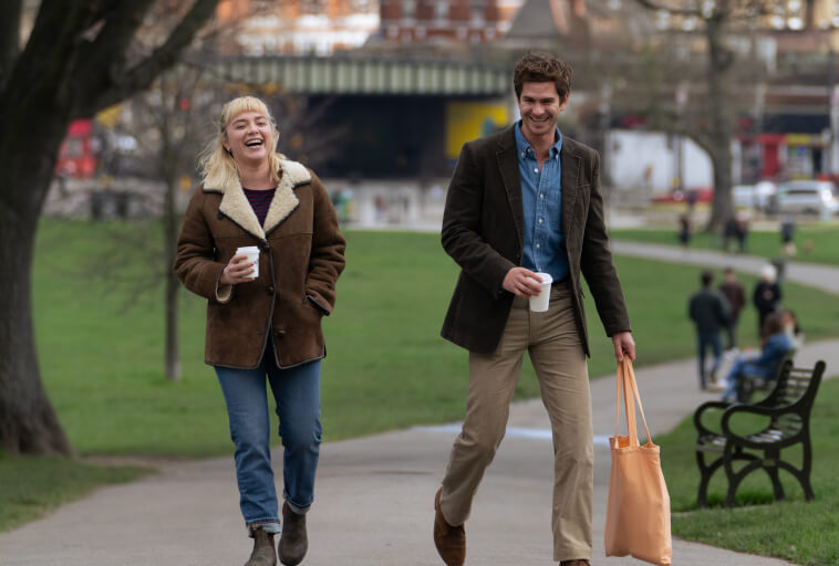 Dwie osoby spacerują razem w parku, uśmiechając się i trzymając kubki z kawą. Osoba po lewej stronie ma krótkie blond włosy i brązową kurtkę; osoba po prawej stronie nosi marynarkę i pomarańczową torbę.