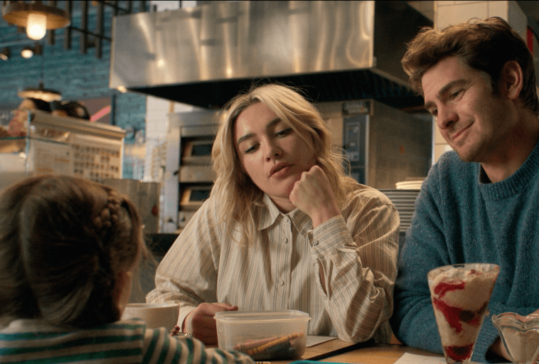 Kobieta i mężczyzna siedzą przy stoliku w przytulnej knajpce, patrząc na małe dziecko naprzeciwko nich. Kobieta wygląda na zamyśloną, opierając podbródek na dłoni. Na stole znajduje się deser i przekąski.