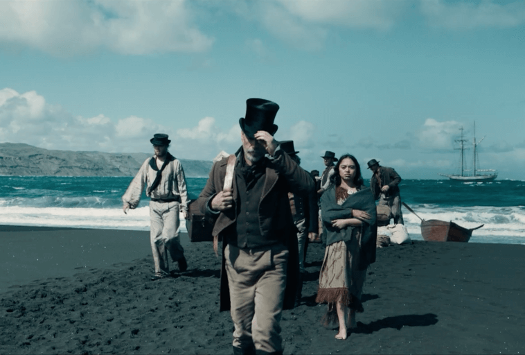 Grupa ludzi w staromodnych ubraniach idzie wzdłuż ciemnej piaszczystej plaży. Mężczyzna z przodu poprawia kapelusz, a młoda kobieta w podartej sukience przygląda się temu. Za nimi widać ocean i wysoki statek.