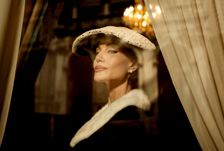 Kobieta w kapeluszu z szerokim rondem i białym futrzanym płaszczu spogląda pewnie w stronę kamery, stojąc między eleganckimi zasłonami z żyrandolem świecącym delikatnie w tle.