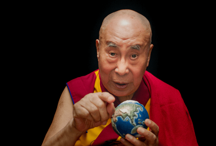 Starszy mężczyzna w tradycyjnym czerwono-żółtym ubraniu wskazuje na małą kulę ziemską, którą trzyma, z poważnym wyrazem twarzy, na prostym czarnym tle.