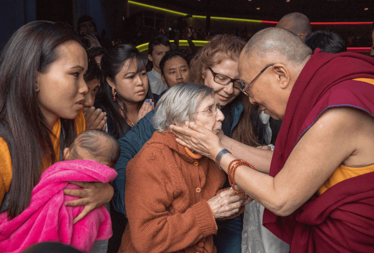 Buddyjski mnich delikatnie trzyma starszą kobietę za twarz, otoczoną przez grupę ludzi, w tym kobietę trzymającą dziecko owinięte w różowy koc. Scena jest ciepła i pełna współczucia.