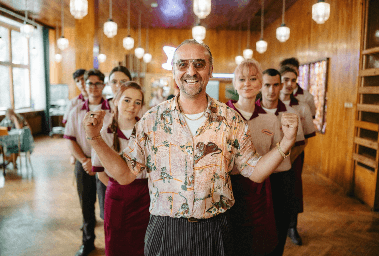 Uśmiechnięty mężczyzna we wzorzystej koszuli stoi przed grupą młodych ludzi ubranych w dopasowane uniformy w jasno oświetlonym pomieszczeniu wyłożonym drewnianymi panelami, co sugeruje zespół pracowników restauracji lub kawiarni.