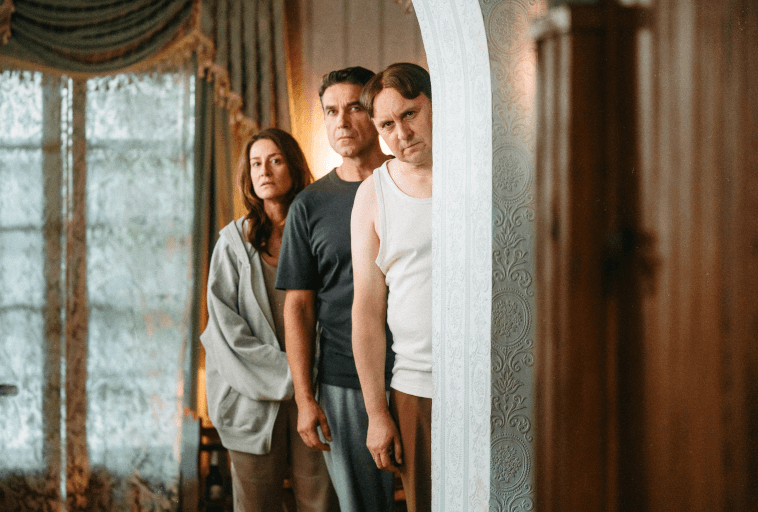 Trzy osoby stoją blisko siebie w pomieszczeniu, wpatrując się uważnie w ozdobną, białą ściankę działową. Kobieta stoi z przodu, a za nią dwóch mężczyzn. Wygląda na to, że jest to salon z wyszukanymi zasłonami.