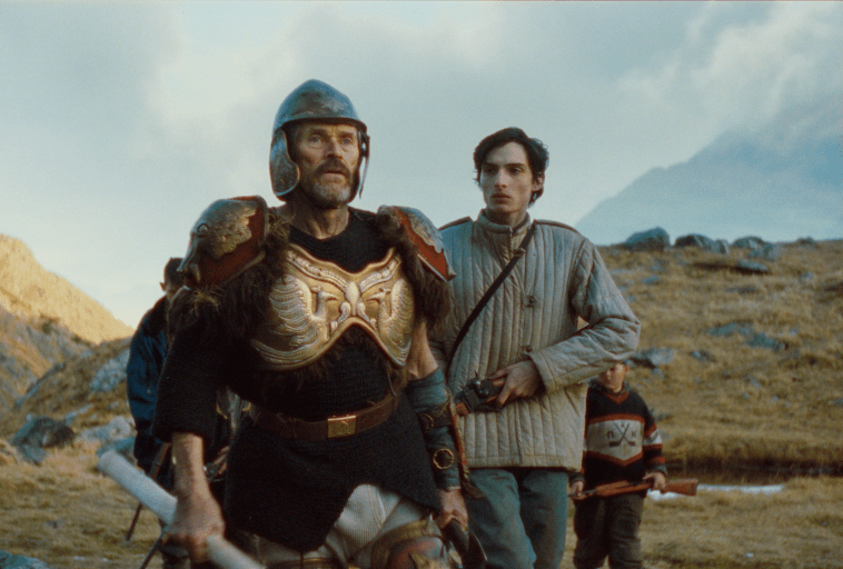 Mężczyzna w ozdobnej, starożytnej zbroi i hełmie stoi z mieczem, podczas gdy dwóch mężczyzn w nowoczesnej odzieży outdoorowej stoi za nim w skalistym, górskim krajobrazie pod zachmurzonym niebem.