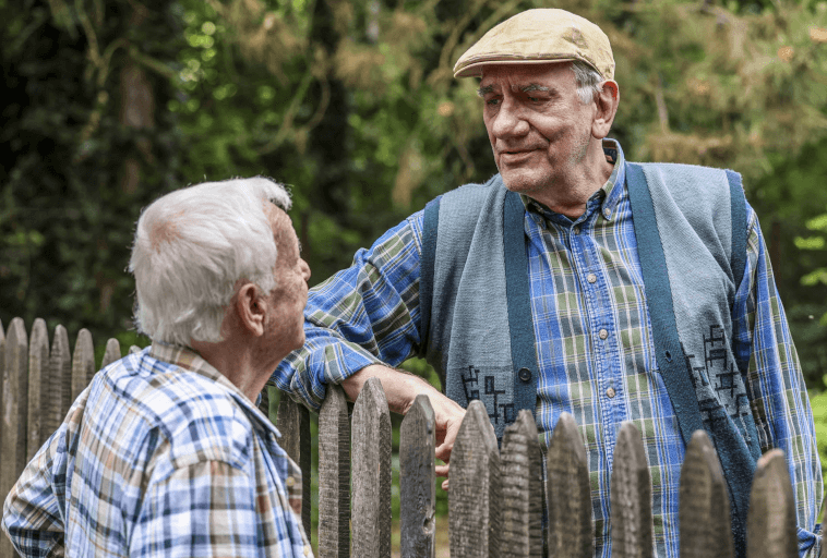 Dwóch starszych mężczyzn w kraciastych koszulach prowadzi przyjacielską rozmowę przy drewnianym płocie na zewnątrz, z zielenią w tle. Jeden z mężczyzn ma na sobie czapkę i kamizelkę, a obaj uśmiechają się do siebie ciepło.