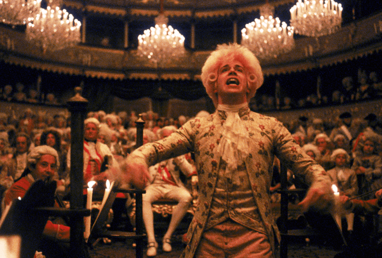 Mężczyzna w ozdobnym XVIII-wiecznym kostiumie i pudrowanej peruce z pasją dyryguje orkiestrą w wielkim, oświetlonym świecami teatrze wypełnionym elegancko ubranymi widzami.