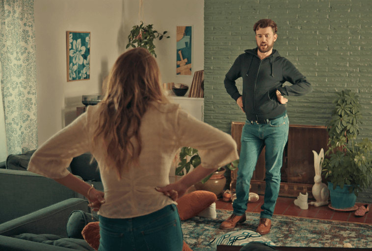 Mężczyzna i kobieta stoją naprzeciwko siebie z rękami na biodrach w salonie. W pokoju znajdują się rośliny, dzieła sztuki na ścianach, kanapa i przedmioty dekoracyjne na podłodze. Atmosfera wydaje się napięta lub konfrontacyjna.