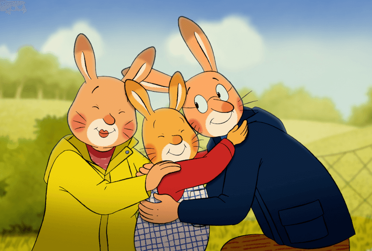 Trzy kreskówkowe króliki, dwoje dorosłych i jedno dziecko, uśmiechają się i przytulają na zewnątrz. Dorośli mają na sobie żółte i niebieskie kurtki, a dziecko nosi czerwoną koszulę. W tle widać zielone pole i drzewa pod błękitnym niebem.