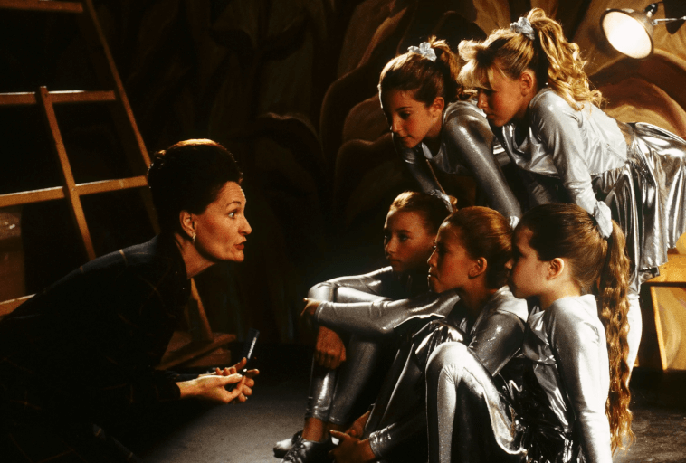 Kobieta przykuca i mówi do pięciu młodych dziewcząt ubranych w błyszczące srebrne kostiumy, które siedzą i uważnie słuchają za kulisami teatru.