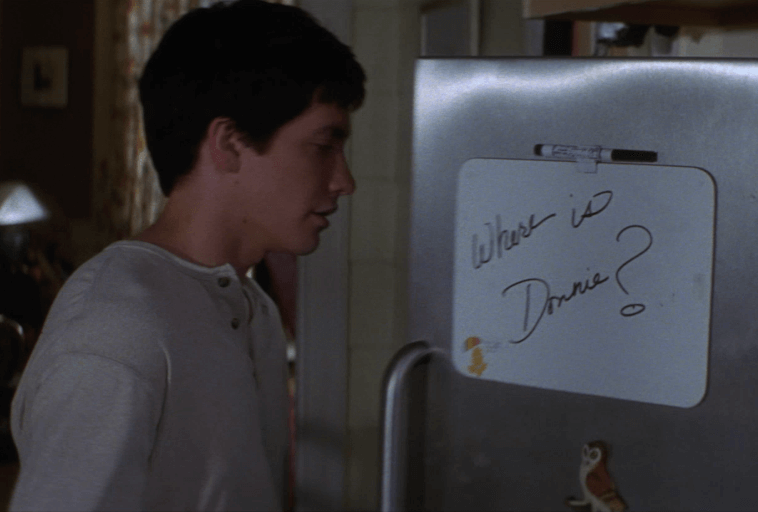 Młoda osoba stoi obok lodówki z przymocowaną tablicą. Na tablicy jest napisana markerem wiadomość: Gdzie jest Donnie? ze znakiem zapytania.
