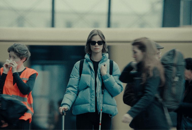 Kobieta w okularach przeciwsłonecznych i niebieskiej kurtce puchowej idzie z białą laską, co wskazuje na upośledzenie wzroku. Jest otoczona przez ludzi, w tym osobę w pomarańczowej kamizelce, w plenerze miejskim.