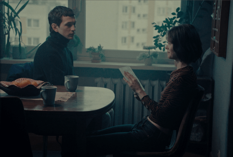 Dwie osoby siedzą naprzeciwko siebie przy stole w słabo oświetlonym pokoju z roślinami domowymi i oknem. Jedna osoba czyta kartkę papieru, podczas gdy druga patrzy; na stole stoją dwie filiżanki.