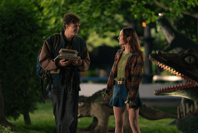 Młody mężczyzna z książkami i plecakiem uśmiecha się do młodej kobiety w kraciastej kurtce i spódnicy. Stoją na zewnątrz w nocy przy posągach dinozaurów pod zielonymi drzewami.