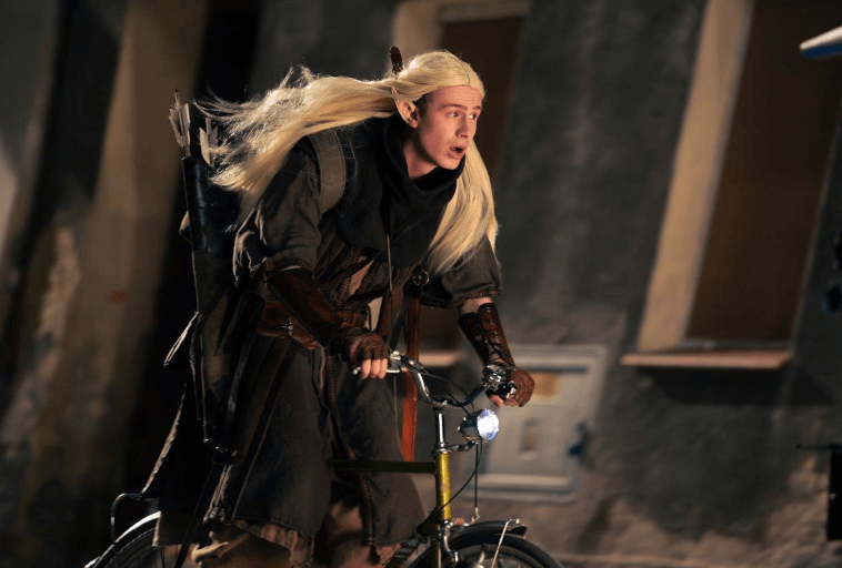 Osoba przebrana za elfa fantasy z długimi blond włosami, ubrana w ciemny płaszcz i nosząca kołczan strzał, jedzie rowerem po słabo oświetlonej ulicy.