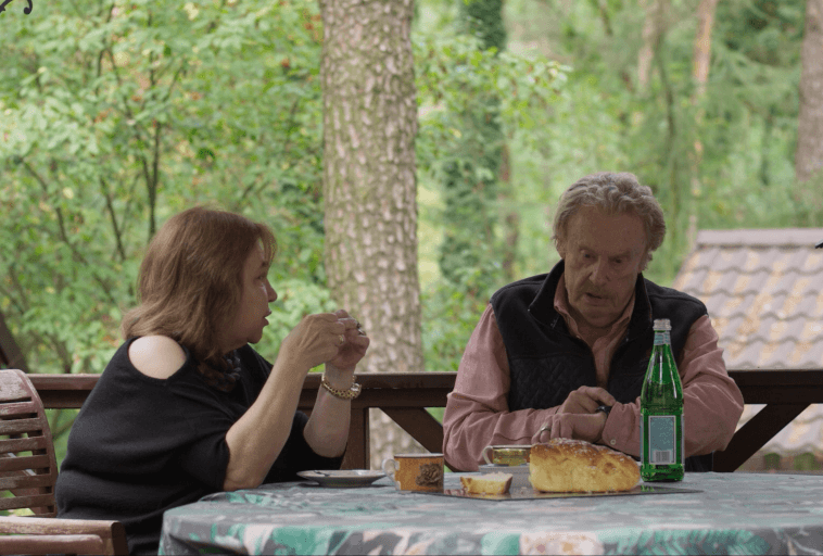 Starszy mężczyzna i kobieta siedzą przy stole na świeżym powietrzu w zalesionej okolicy, rozmawiając przy posiłku z chlebem, filiżankami i zieloną szklaną butelką przed sobą. Stół przykryty jest wzorzystym obrusem.