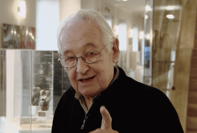 Starszy mężczyzna z białymi włosami i okularami, ubrany w ciemny sweter zapinany na zamek, uśmiecha się i gestykuluje prawą ręką w dobrze oświetlonym wnętrzu z gablotami i dziełami sztuki w tle.