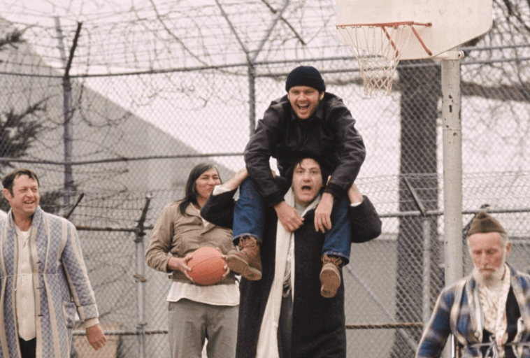 Czterech mężczyzn gra w koszykówkę na boisku zewnętrznym; jeden siedzi na ramieniu drugiego, uśmiechając się. Dwóch innych, z których jeden trzyma piłkę do koszykówki, przygląda się rozgrywce. Ogrodzenie z drutu kolczastego otacza boisko.