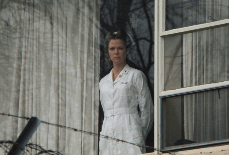 Kobieta w białym uniformie pielęgniarskim stoi za szklanym oknem z zasłonami, patrząc na zewnątrz z poważnym wyrazem twarzy. Na pierwszym planie widoczne są bezlistne gałęzie drzew i ogrodzenie.