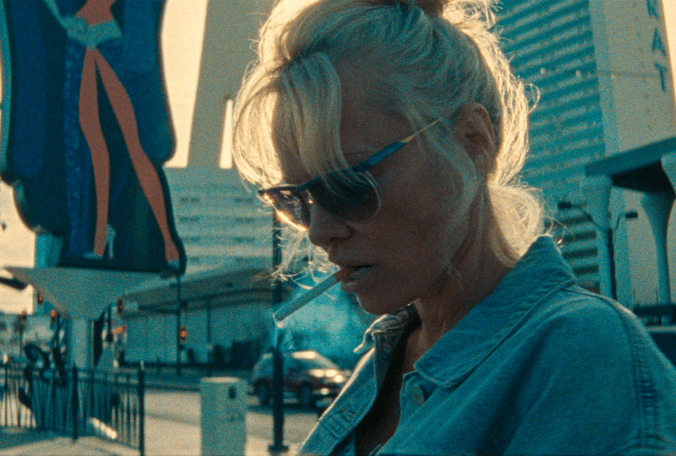 Kobieta z blond włosami w niechlujnym kucyku, w okularach przeciwsłonecznych i dżinsowej kurtce, stoi na zewnątrz z papierosem w ustach. Tło przedstawia miejskie budynki i znaki uliczne w miękkiej ostrości.