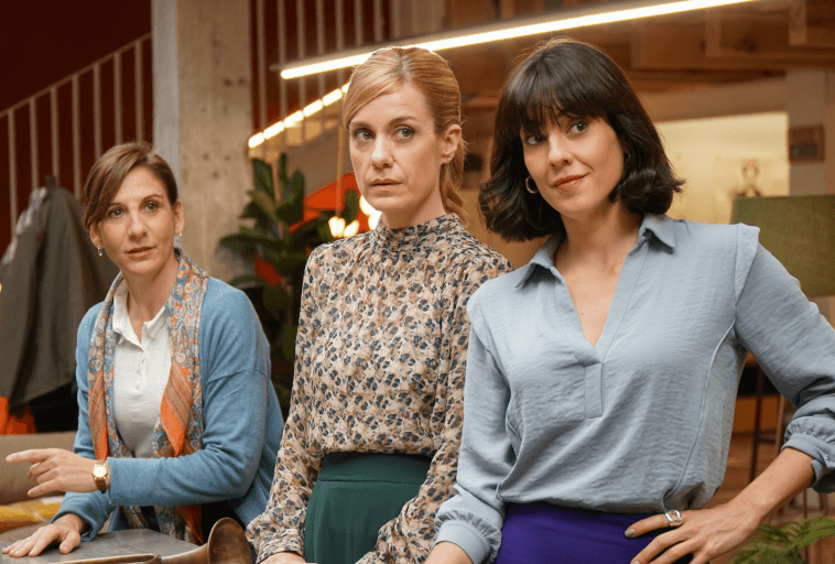 Trzy kobiety stoją w pomieszczeniu, patrząc w stronę kamery. Mają poważne lub zamyślone miny. Tło obejmuje nowoczesne meble, ciepłe oświetlenie i schody.