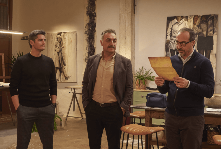 Trzech mężczyzn stoi w pokoju ze sztuką i roślinami; jeden mężczyzna po prawej czyta z gazety, podczas gdy pozostali dwaj słuchają. Otoczenie wydaje się swobodne i kreatywne, z obrazami i stołkami w tle.