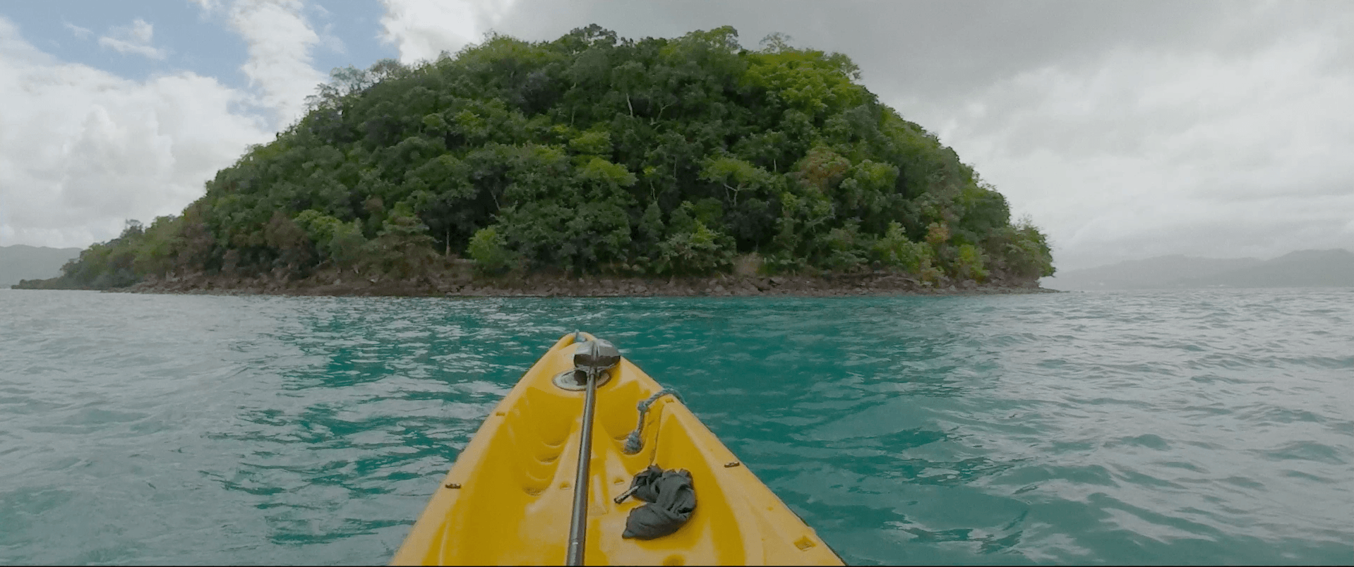 Żółty kajak zbliża się do małej, bujnej zielonej wyspy otoczonej turkusową wodą pod zachmurzonym niebem.