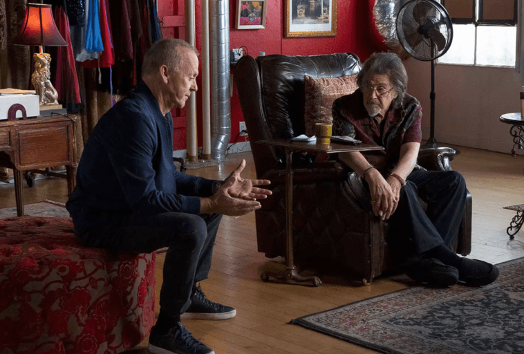 Dwóch starszych mężczyzn siedzi w ciepło oświetlonym, eklektycznym pokoju; jeden na czerwonej otomanie, gestykulując podczas rozmowy, drugi w skórzanym fotelu, słuchając uważnie z brodą w dłoni.