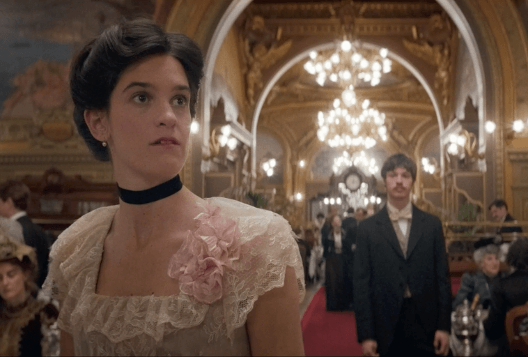 Młoda kobieta w kremowej sukience w stylu vintage z różowym kwiatem stoi w ozdobnej, oświetlonej żyrandolem sali balowej. Za nią elegancko ubrani ludzie mieszają się, a mężczyzna w smokingu patrzy w jej kierunku.
