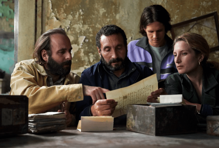 Cztery osoby siedzą przy stole i analizują stare papiery i dokumenty w słabo oświetlonym, zużytym pomieszczeniu. Jeden z mężczyzn wskazuje na list, podczas gdy pozostali przyglądają mu się uważnie, sugerując poważną dyskusję lub dochodzenie.