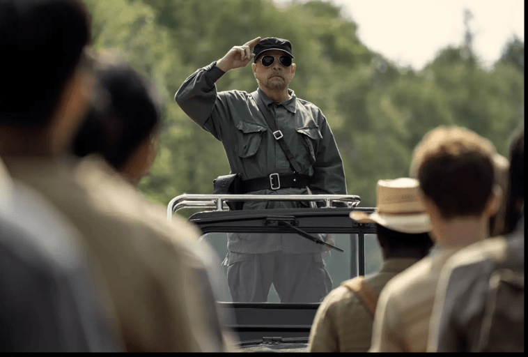 Mężczyzna w mundurze w stylu wojskowym i okularach przeciwsłonecznych salutuje, stojąc w otwartym pojeździe, twarzą do grupy ludzi na zewnątrz, z drzewami w tle.