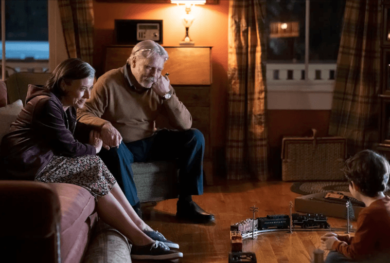 Starszy mężczyzna i kobieta siedzą na kanapie, uśmiechając się i obserwując małego chłopca bawiącego się zabawkowym pociągiem na drewnianej podłodze w przytulnym, ciepło oświetlonym salonie.