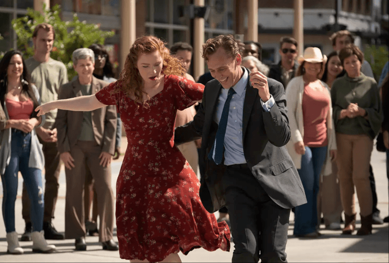 Kobieta w czerwonej sukience w kwiaty i mężczyzna w garniturze tańczą energicznie na świeżym powietrzu, otoczeni przez wesoły tłum obserwujący i uśmiechający się w tle.