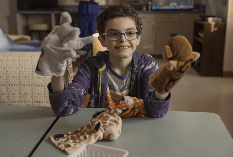 Uśmiechnięte dziecko w okularach bawi się pacynkami zwierząt przy stole, trzymając po jednej pacynce w każdej ręce i więcej pacynek spoczywających na stole przed nim.