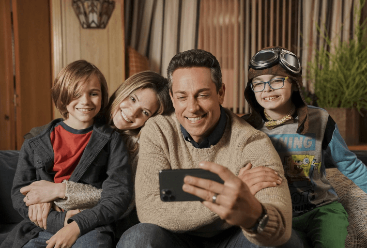 Uśmiechnięta czteroosobowa rodzina siedzi na kanapie i robi sobie selfie. Ojciec trzyma telefon, matka opiera się na jego ramieniu, a dwoje dzieci, jedno w okularach lotniczych, siedzi blisko siebie, wszyscy wyglądają na szczęśliwych i zrelaksowanych.