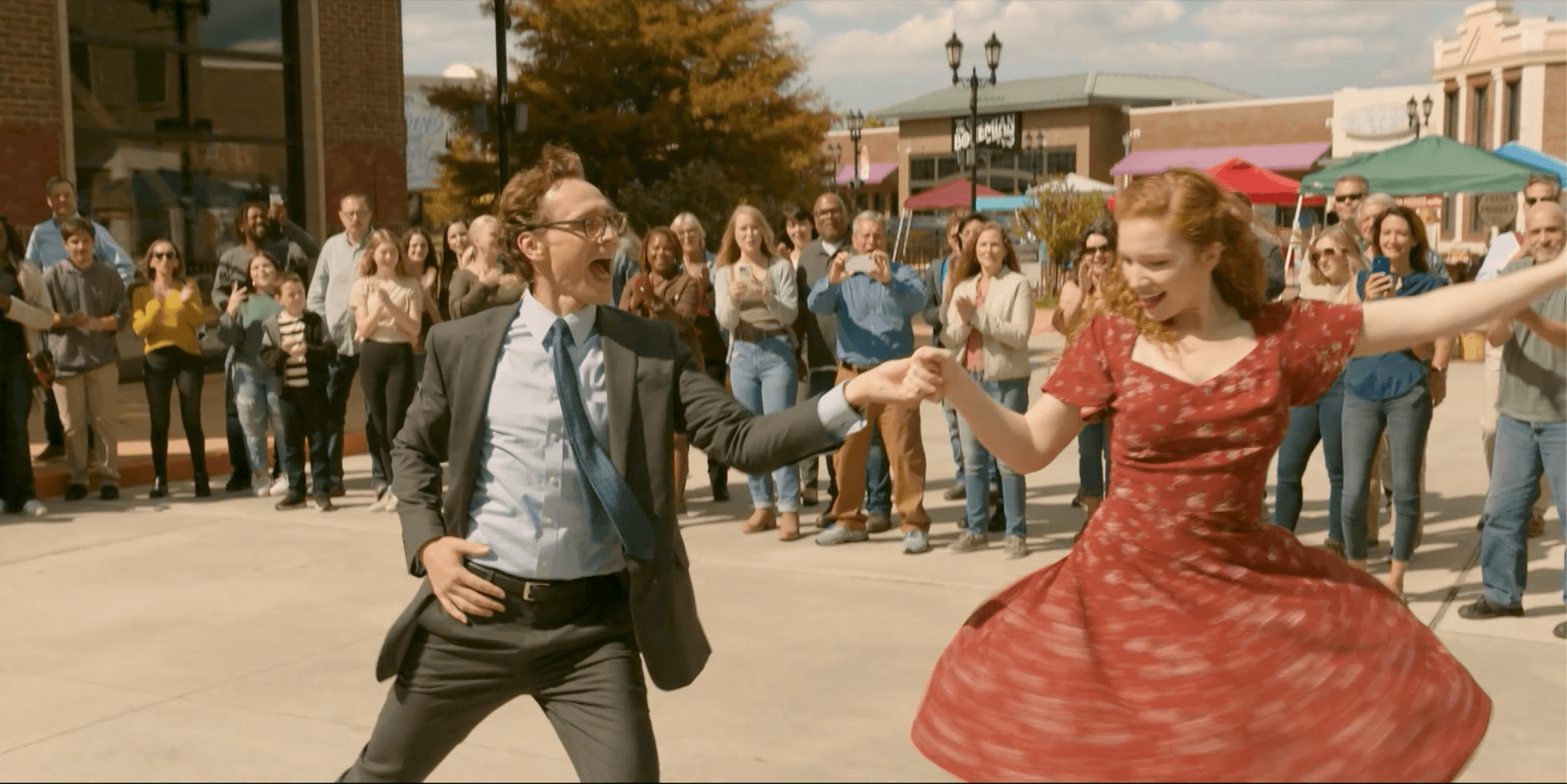 Mężczyzna w garniturze i kobieta w czerwonej sukience energicznie tańczą razem na świeżym powietrzu, otoczeni wiwatującym tłumem ludzi w słoneczny dzień.