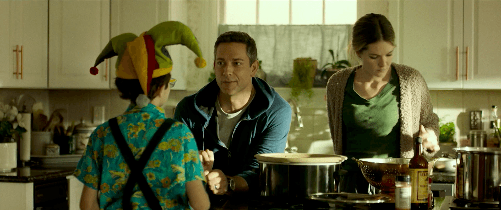 Mężczyzna siedzi przy blacie kuchennym, patrząc na dziecko w błazeńskim kapeluszu i kolorowej koszuli, podczas gdy kobieta w zielonej bluzce gotuje przy kuchence w ciepło oświetlonej kuchni.