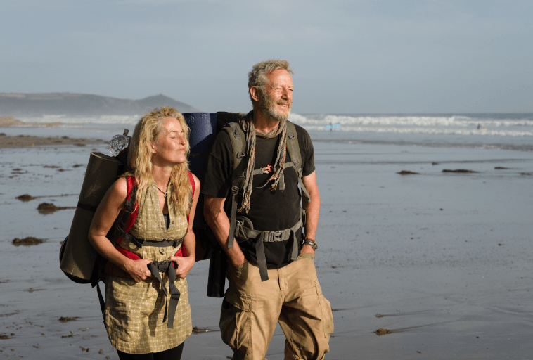 Starsza para z plecakami stoi na piaszczystej plaży, uśmiechnięta i zwrócona w stronę słońca. Za nimi przetaczają się fale oceanu, a pod czystym niebem widać odległe wzgórza.